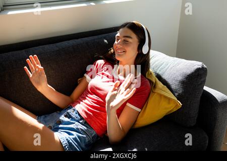 Das kaukasische Teenager-Mädchen entspannt sich zu Hause, hört Musik auf ihren hübschen Kopfhörern und sonnt sich im Sonnenlicht. Stockfoto