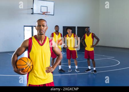Ein afroamerikanischer Mann hält einen Basketball in einem Hallenplatz und verkörpert einen sportlichen Lebensstil. Teamkollegen stehen hinter ihm bereit und heben Teamwork A hervor Stockfoto