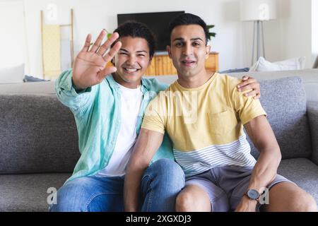 Ein vielseitiges schwules Paar genießt einen entspannten Videoanruf zu Hause. Sie teilen sich eine gemütliche Couch, die Glück und Gesellschaft ausstrahlt. Stockfoto