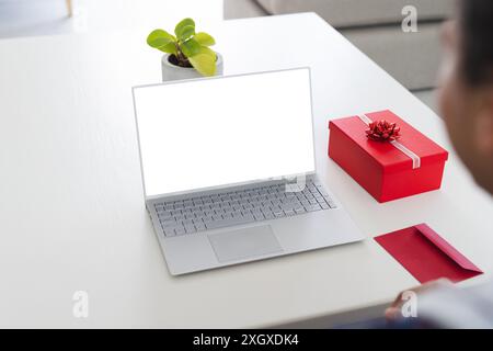 Der Laptop-Bildschirm zeigt auf einem weißen Schreibtisch zur Kamera und verfügt über einen Kopierbereich. Eine rote Geschenkbox verleiht dem Büro einen Hauch von Feiern. Stockfoto