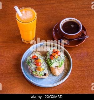 Teller mit zwei Avocadotoasts, einem Glas frischen Orangensaft und einer Tasse schwarzen Kaffee auf Holztisch Stockfoto