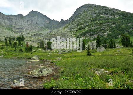 Große Steine auf der Oberfläche eines wunderschönen Sees und eine malerische Lichtung umgeben von hohen Klippen an einem bewölkten Sommertag. Marmor Lake, Ergaki Natur Stockfoto