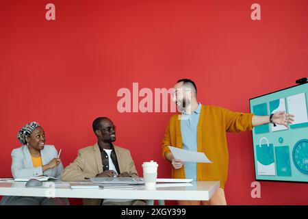 Mann, der mit dem Team eine ansprechende Präsentation in moderner Büroumgebung mit roter Wand und großem Display durchführt. Teammitglieder hören aufmerksam zu und nehmen an Diskussionen Teil Stockfoto