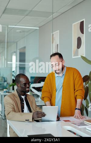 Zwei verschiedene männliche Kollegen, Caucasian man und Black man, lächeln, während sie Projektdetails auf einem digitalen Tablet in modernen Büros diskutieren Stockfoto