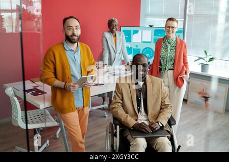 Gruppe verschiedener Kollegen, die sich in einem modernen Büro mit Präsentationsbildschirm im Hintergrund ausgeben. Schwarzer Mann im Rollstuhl sitzt vorne, während andere lächelnd um den Tisch stehen Stockfoto