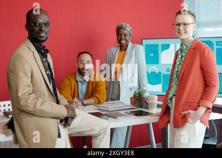Gruppe verschiedener Kollegen in zeitgenössischen Büros. Vier Fachleute posieren am Tisch mit digitalem Bildschirm im Hintergrund und fördern so die Zusammenarbeit und Kreativität Stockfoto