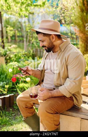 Glücklicher bärtiger männlicher Bauer mit Handy, der Online-Videos ansieht oder im Videochat kommuniziert, während er im Garten sitzt Stockfoto