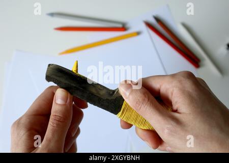 Foto des Verfahrens des Schärfens von Farbstiften mit einem Klerikmesser. Menschliche Hände. Gelber Bleistift. Metallmesser. Im Hintergrund stehen Weißbücher und Stockfoto