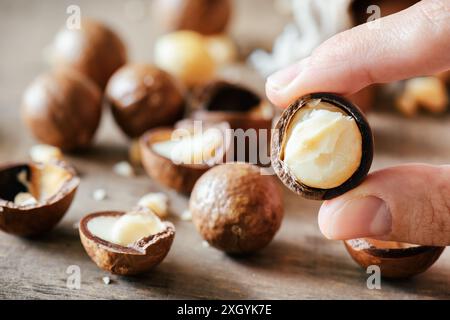 Nahaufnahme von Fingern, die Macadamia-Nuss halten. Macadamia-Nüsse auf Holzbrett im Hintergrund. Stockfoto