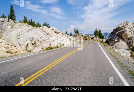 Eine zweispurige Straße mit doppelten gelben Linien windet sich zwischen verwirrt liegenden weißen Felsbrocken hin zu Kiefern bei den Hoodoos im Yellowstone-Nationalpark, Wyoming. Stockfoto