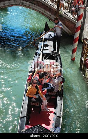 Eine Gruppe von sechs jungen asiatischen Touristen saß bei einer Gondelfahrt und wurde an einem sonnigen Tag in Venedig an einer Brücke entlang eines Kanals vorbei gerudert Stockfoto