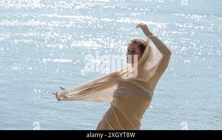 Eine Frau posiert anmutig am glitzernden Wasser, bedeckt mit einem leichten, durchscheinenden Stoff. Das Sonnenlicht glitzert auf den Wellen und schafft ein ruhiges und ruhiges Ambiente Stockfoto