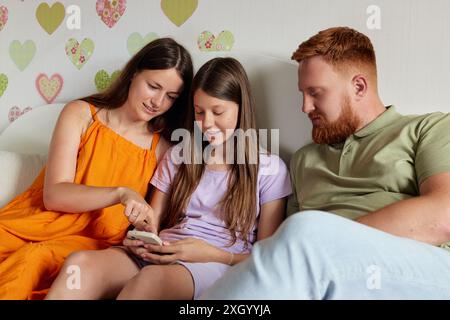 Mutter, Vater und Tochter sehen sich gemeinsam ein Smartphone auf einer weißen Couch an Stockfoto