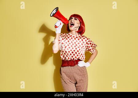 Eine selbstbewusste rothaarige Frau im Pop-Art-Make-up hält ein rot-weißes Megaphon auf gelbem Hintergrund. Stockfoto