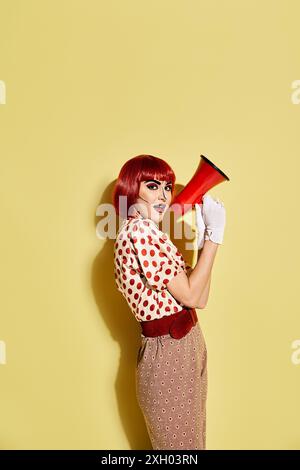 Eine lebendige rothaarige Frau mit Pop-Art-Make-up hält ein rot-schwarzes Megaphon vor einem hellgelben Hintergrund. Stockfoto