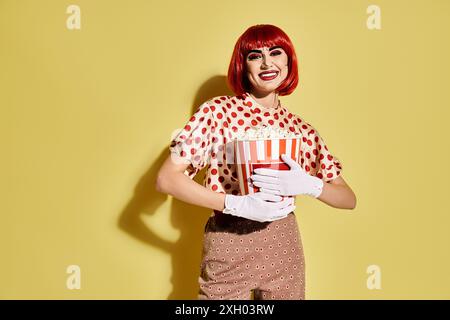 Eine hübsche rothaarige Frau in Polka Dot-Bluse mit Pop-Art-Make-up, die eine Kiste Popcorn auf gelbem Hintergrund hält. Stockfoto