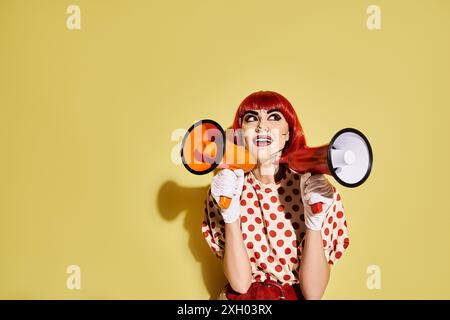 Eine auffällige rothaarige Frau mit Pop-Art-Make-up hält ein Megaphon vor einem leuchtend gelben Hintergrund. Stockfoto