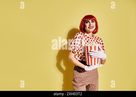 Eine hübsche rothaarige Frau mit Pop-Art-Make-up hält eine Kiste Popcorn vor einem leuchtend gelben Hintergrund. Stockfoto