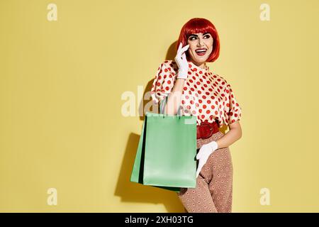 Eine lebendige rothaarige Frau mit Pop-Art-Make-up hält eine grüne Tasche, während sie auf einem Handy vor gelbem Hintergrund chatten. Stockfoto