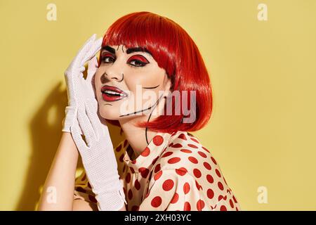 Eine auffällige Rothaarige mit Pop-Art-Make-up trägt ein Kleid mit Punktmuster und elegante weiße Handschuhe vor leuchtendem gelbem Hintergrund. Stockfoto