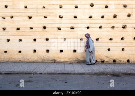 Komposition einer älteren Frau, die in einer grauen Djellaba gekleidet vor der massiven Stadtmauer von Fès in Marokko, Nordafrika, die Straße hinuntergeht Stockfoto