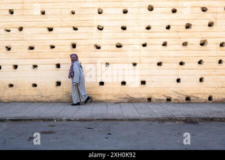 Komposition einer älteren Frau, die in einer grauen Djellaba gekleidet vor der massiven Stadtmauer von Fès in Marokko, Nordafrika, die Straße hinuntergeht Stockfoto