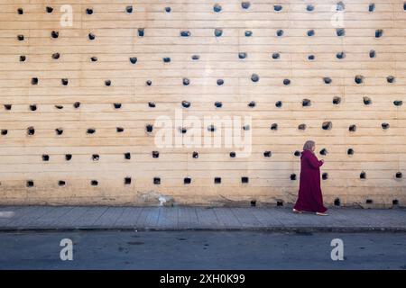 Komposition einer Frau, die in einer burgunderroten Djellaba vor der massiven Stadtmauer von Fès in Marokko, Nordafrika, die Straße entlang geht Stockfoto