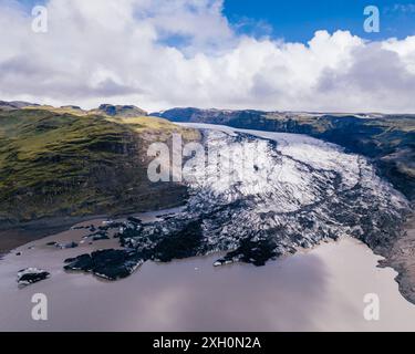 Blick aus der Vogelperspektive auf den Sólheimajokull-Gletscher in Island, mit kontrastierenden grünen Hügeln und dem eisigen, zerklüfteten Gletscher, der auf eine Gletscherlagune unter einem partl trifft Stockfoto