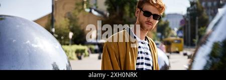 Junger rothaariger Mann, der in gelber Strickjacke und Sonnenbrille debonair aussieht, während er durch die Stadt schlendert. Stockfoto