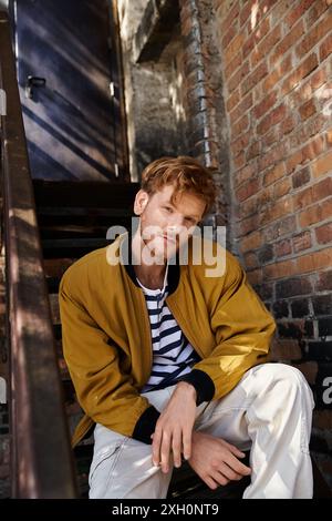 Ein junger Mann mit rotem Haar in stilvoller Kleidung sitzt auf der Bautreppe. Stockfoto