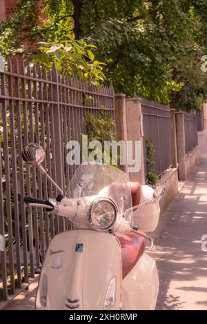 Eine weiße Vespa, die in einer sonnendurchfluteten Straße neben einem Zaun und Bäumen parkt, Mailand, Italien Stockfoto