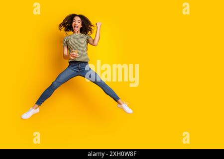 Foto in voller Größe des überfröhlichen funky Mädchen gekleidet T-Shirt halten Smartphone springend Gewinn Lotterie schreien isoliert auf gelbem farbigen Hintergrund Stockfoto