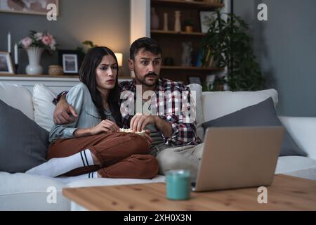 Ein Paar schockiert, sich Horrorfilm auf einem Laptop anzusehen, der zu Hause auf dem Sofa sitzt und Popcorn isst. Angst in ihren Gesichtern, gruselige Filme und Wochenendaktivitäten Stockfoto