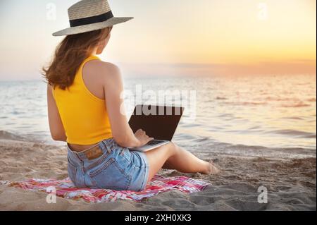 Rückansicht Junge kaukasische Frau in Shorts und Strohhut benutzt ihren Laptop, während sie bei Sonnenuntergang am Sandstrand am Meer sitzt. Freiberufler und Stockfoto