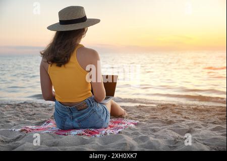 Rückansicht Junge kaukasische Frau in Shorts und Strohhut benutzt ihren Laptop, während sie bei Sonnenuntergang am Sandstrand am Meer sitzt. Freiberufler und Stockfoto