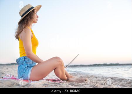 Nette junge Frau mit weißem Laptop am Strand am Abend. Freiberufler arbeiten Remote über das Internet Stockfoto