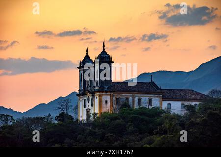 Alte und historische Kirche auf dem Hügel während des Sonnenuntergangs in der Stadt Ouro Preto in Minas Gerais, Brasilien mit den Bergen hinter Stockfoto