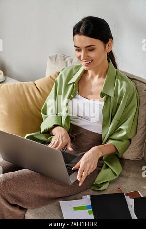 Eine Frau in lässiger Kleidung arbeitet ferngesteuert auf einem Laptop, während sie auf einer Couch sitzt. Stockfoto