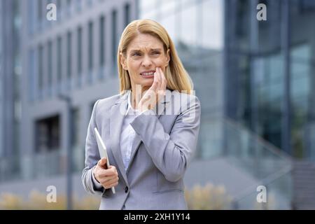 Geschäftsfrau in formeller Kleidung, die Schmerzen hat, die Wange vor dem modernen Bürogebäude berührt. Zeigt Beschwerden, Zahnschmerzen, Stress im professionellen Umfeld. Stockfoto