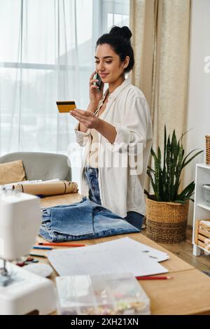 Eine junge Frau in lässiger Kleidung bringt ihre Kleidung auf, während sie auf einem Tisch sitzt und ein Handy benutzt. Stockfoto