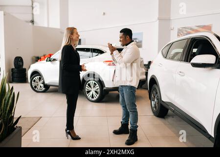 Seitliche Aufnahme eines glücklichen afrikanischen Autohauses, der den Schlüssel von einer lächelnden Autohändlerin im Händlerbetrieb erhält. Fröhlicher schwarzer Mann, der den Schlüssel zum gekauften Auto nimmt und Stockfoto