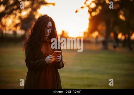 Eine Frau mit langen lockigen Haaren und einer Brille überprüft ihr Telefon, während sie im Park steht und im warmen Licht der untergehenden Sonne gebadet ist. Stockfoto