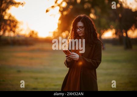 Eine Frau mit langen, lockigen Haaren, die einen braunen Mantel und ein rostfarbenes Kleid trägt, benutzt ihr Smartphone, während sie in einem Park mit untergehender Sonne steht. Stockfoto