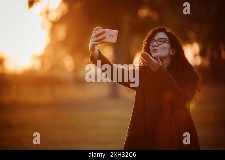 Eine Frau in Brille macht ein Selfie mit einem Handy, während sie einen Kuss bläst, mit der untergehenden Sonne hinter sich. Stockfoto