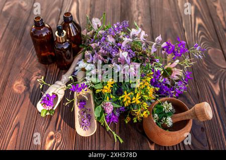 Eine Holzplatte ist mit einem Strauß frischer Kräuter bedeckt, darunter violette Blüten, gelbe Blüten und grüne Blätter. Drei Glasflaschen gefüllt mit Stockfoto