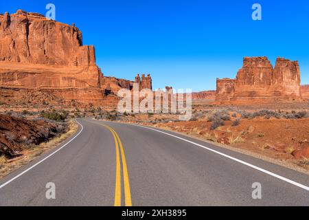 Desert Road – Ein sonniger Blick auf eine Wüstenstraße, die sich in Richtung der hohen roten Sandsteinformationen im Arches National Park, Utah, USA, windet. Stockfoto