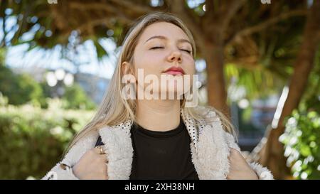Eine junge blonde Frau genießt einen ruhigen Moment in einem üppig grünen Park mit geschlossenen Augen und Sonnenlicht, das durchströmt. Stockfoto