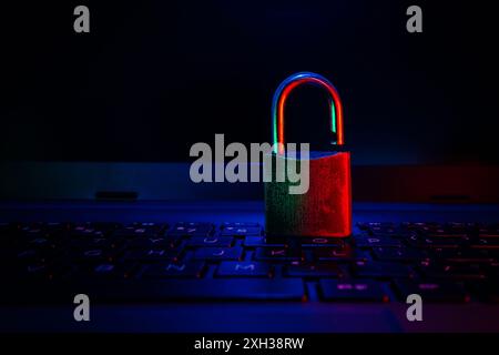 Ein Vorhängeschloss aus Metall befindet sich in einem dunklen Raum auf einer Laptop-Tastatur, die von rotem und blauem Licht beleuchtet wird. Stockfoto