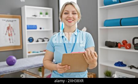 Lächelnde Frau in blauer Uniform mit Klemmbrett in einer gut ausgestatteten Rehabilitationsklinik. Stockfoto