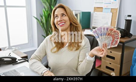 Lächelnde junge Frau, die kolumbianische Pesos in einer modernen Büroumgebung hält Stockfoto